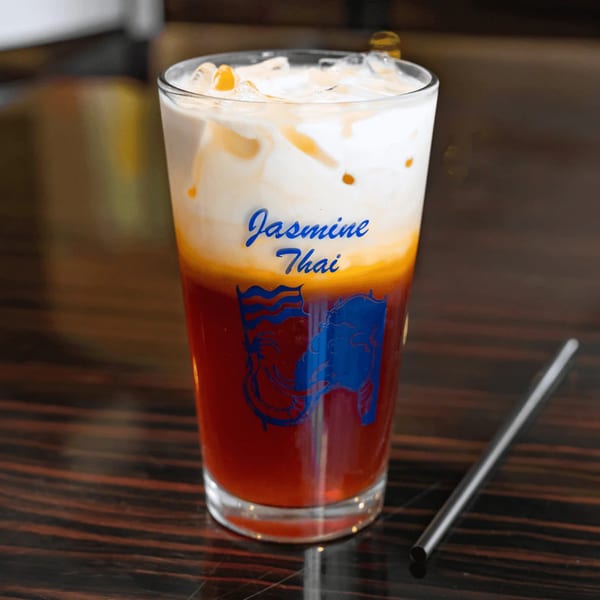 Iced Thai tea with cream and ice in a glass with "Jasmine Thai" logo, next to a metal straw.