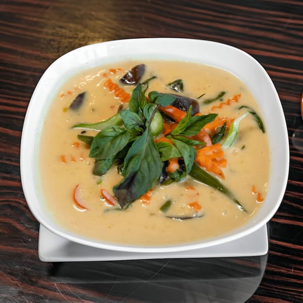 A white bowl of creamy Thai curry with vegetables and fresh basil on a dark wooden table.