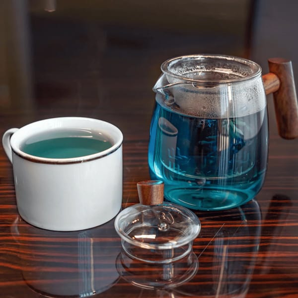 A white mug and a glass teapot filled with vibrant blue butterfly pea tea on a wooden table.